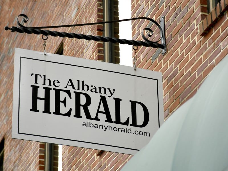 A hanging white sign with bold black text that reads "The Albany Herald" and below it, "albanyherald.com." The sign is mounted on a red brick wall with a black wrought-iron bracket. A light-colored awning partially covers the right side of the image.